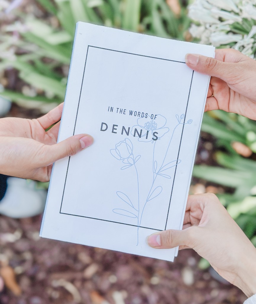Hands holding a Quillegacy memory booklet titled "In the Words of Dennis" outdoors.