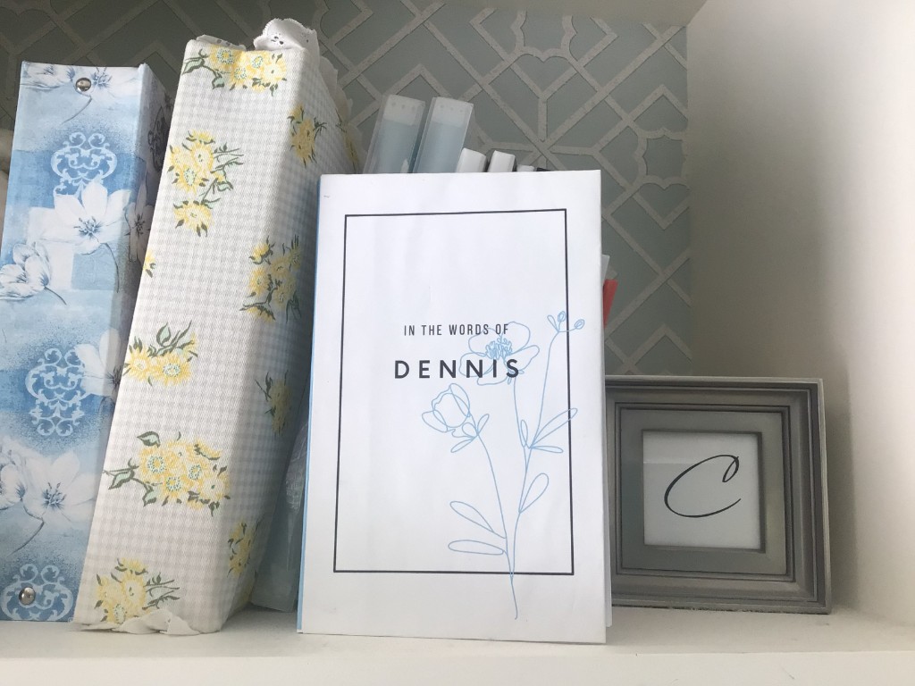 A Quillegacy memory booklet titled "In the Words of Dennis", sitting on a shelf beside floral-patterned books.